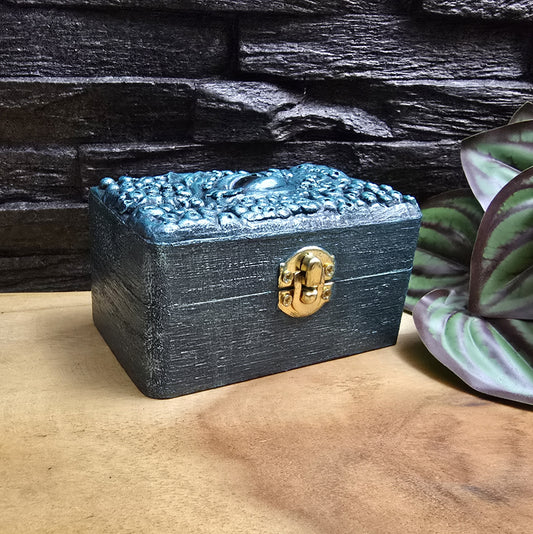 Das Bild zeigt eine geheimnisvolle Schatulle Auge des Oktopus, Meeresgrün, mit Samtkissen und Schnappverschluss. Ladetruhe, Würfelbox, Schmuckkiste
Ob als Schmuckschatulle, Ladebox, Würfelbox oder für andere Kostbarkeiten – diese einzigartige Truhe ist nicht nur ein praktisches Behältnis, sondern ein magisches Artefakt, das die Geschichten der Meere in sich trägt.