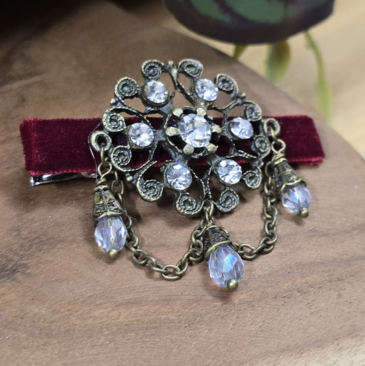 Das Bild zeigt eine Krokodilspange im Vintagelook mit Strass und dunkelrotem Samt, Kristallglasanhänger, Kettchen, Einzelstück
Lass dich von der zeitlosen Eleganz vergangener Epochen verzaubern mit dieser exquisiten Krokodilspange im Vintagestil. Sie ist kunstvoll verziert mit einem filigranen, antik bronzefarbigen Schmuckteil, welches mit funkelndem Strass besetzt ist.