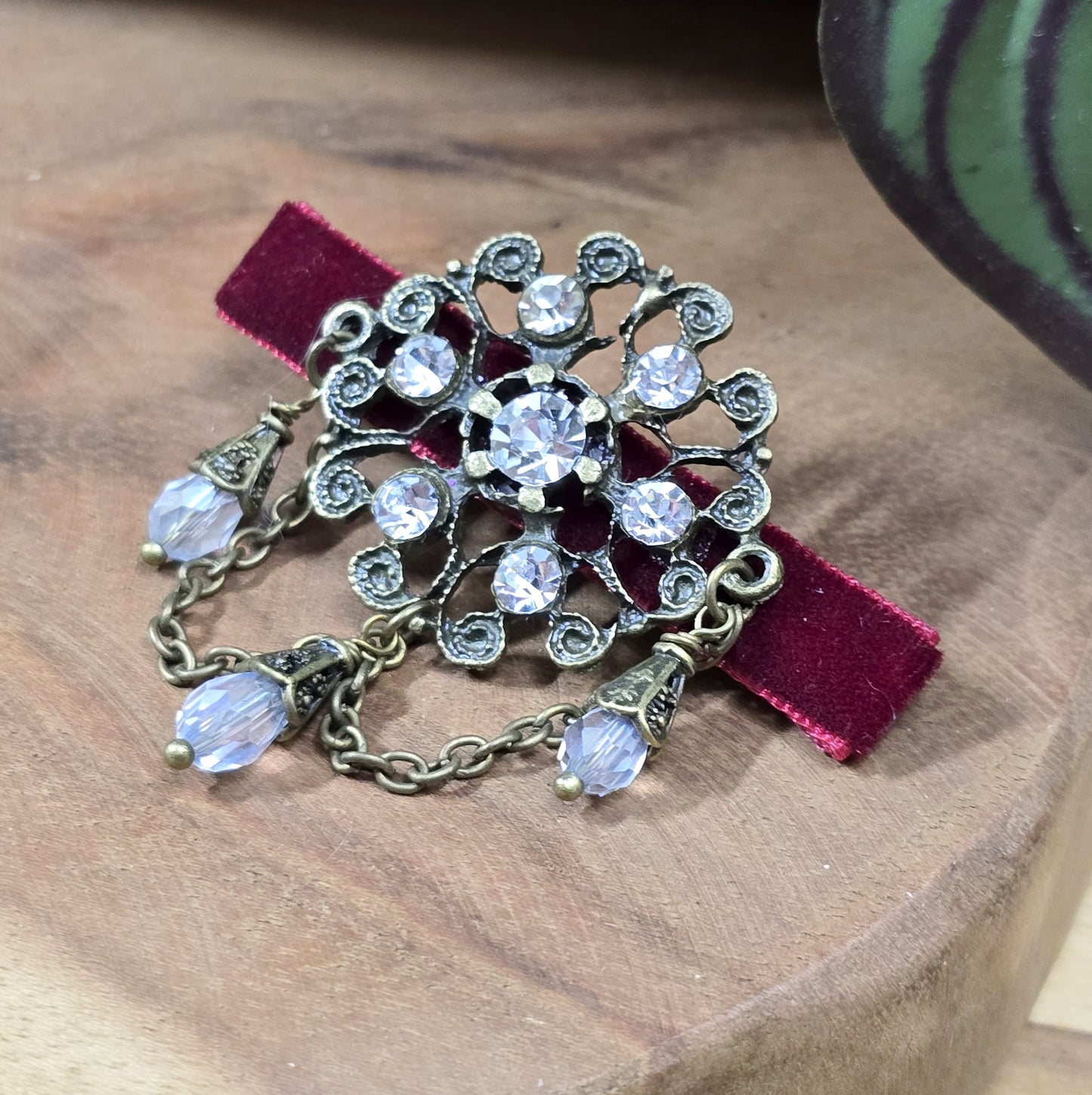 Das Bild zeigt eine Krokodilspange im Vintagelook mit Strass und dunkelrotem Samt, Kristallglasanhänger, Kettchen, Einzelstück
Lass dich von der zeitlosen Eleganz vergangener Epochen verzaubern mit dieser exquisiten Krokodilspange im Vintagestil. Sie ist kunstvoll verziert mit einem filigranen, antik bronzefarbigen Schmuckteil, welches mit funkelndem Strass besetzt ist.