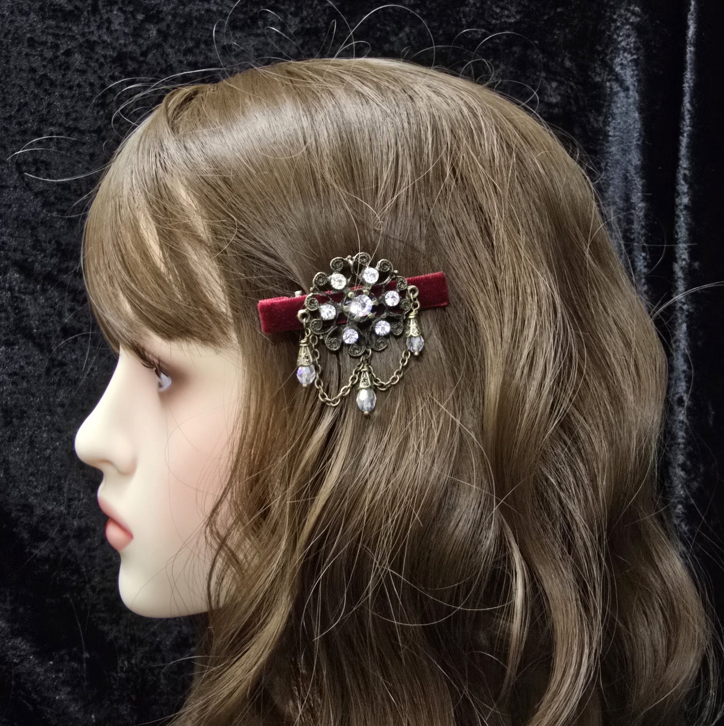 Das Bild zeigt eine Krokodilspange im Vintagelook mit Strass und dunkelrotem Samt, Kristallglasanhänger, Kettchen, Einzelstück
Lass dich von der zeitlosen Eleganz vergangener Epochen verzaubern mit dieser exquisiten Krokodilspange im Vintagestil. Sie ist kunstvoll verziert mit einem filigranen, antik bronzefarbigen Schmuckteil, welches mit funkelndem Strass besetzt ist.