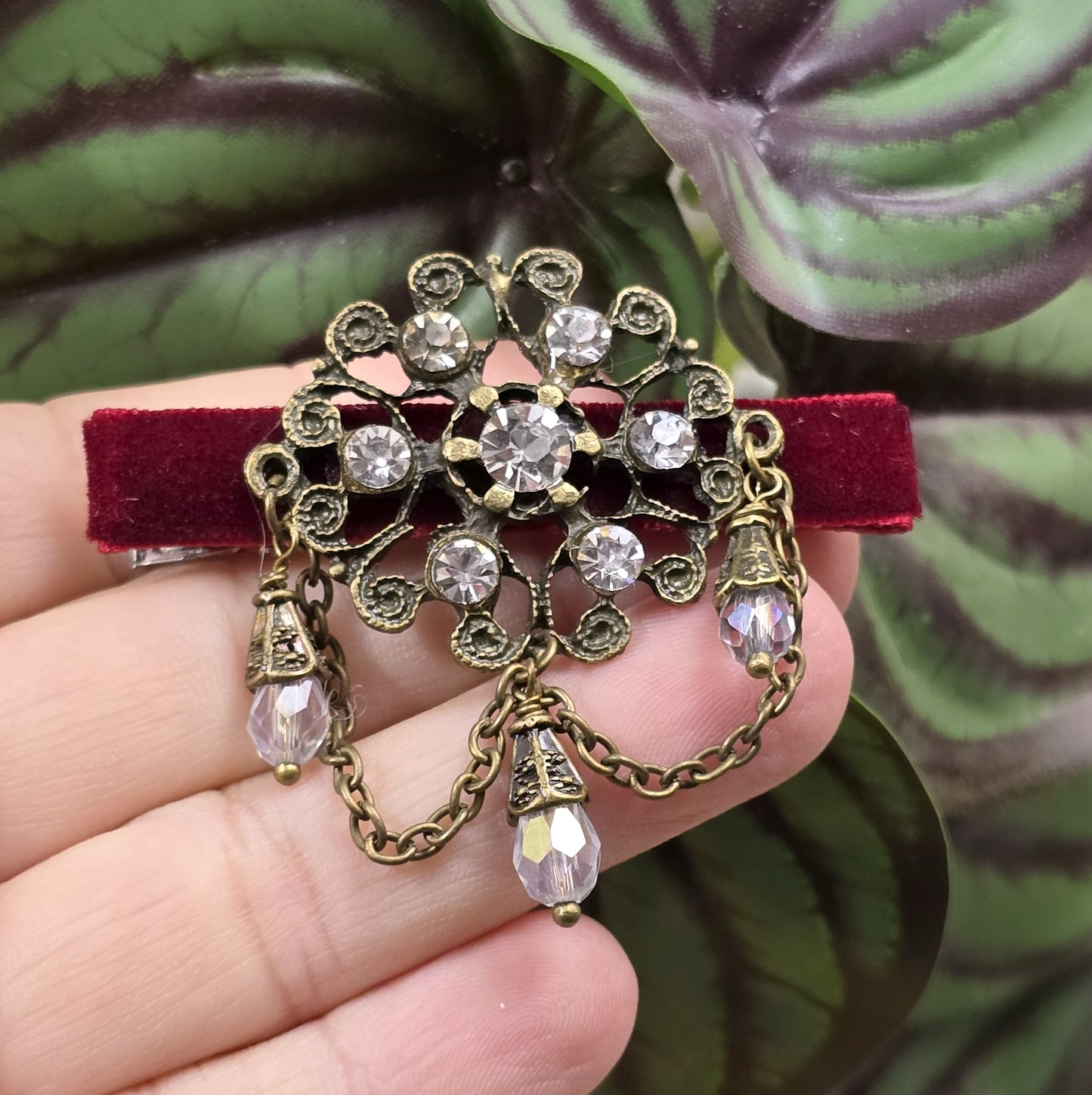 Das Bild zeigt eine Krokodilspange im Vintagelook mit Strass und dunkelrotem Samt, Kristallglasanhänger, Kettchen, Einzelstück
Lass dich von der zeitlosen Eleganz vergangener Epochen verzaubern mit dieser exquisiten Krokodilspange im Vintagestil. Sie ist kunstvoll verziert mit einem filigranen, antik bronzefarbigen Schmuckteil, welches mit funkelndem Strass besetzt ist.