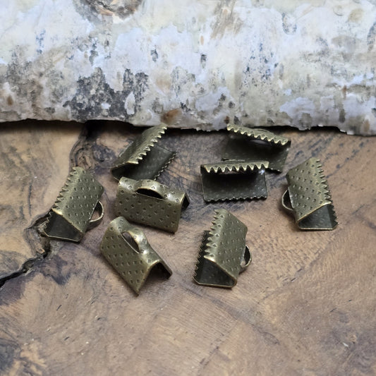 Das Bild zeigt antik bronzefarbige Bandklemmen mit Zähnen, 10 mm breit – ideal zum Befestigen von Verschlüssen an Stoffbändern. Einfach mit einer Zange auf das Bandende klemmen.