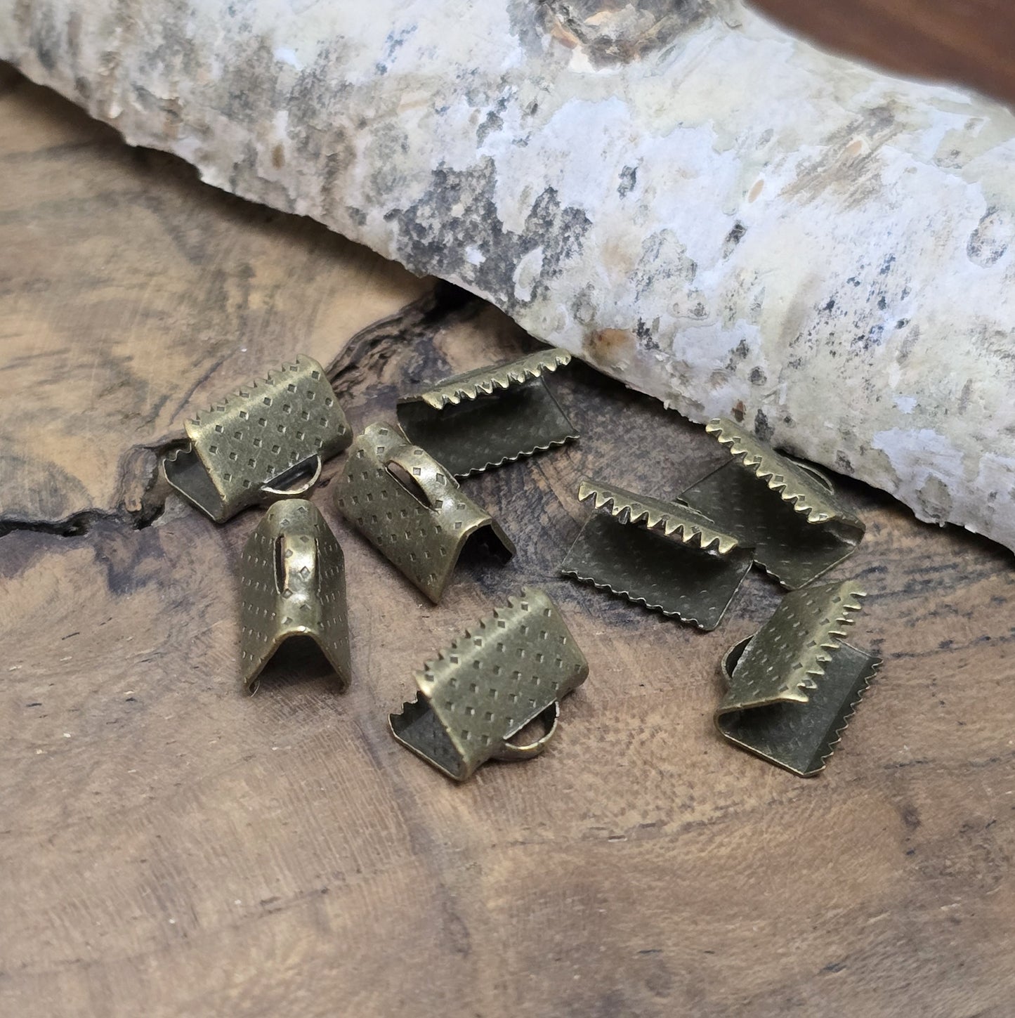 Das Bild zeigt antik bronzefarbige Bandklemmen mit Zähnen, 10 mm breit – ideal zum Befestigen von Verschlüssen an Stoffbändern. Einfach mit einer Zange auf das Bandende klemmen.