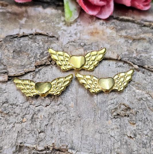 Das Bild zeigt vergoldete Metallperlen in Form von Engelsflügeln mit einem Herz in der Mitte. Die Perlen sind 22 mm groß und eignen sich für die Herstellung von Schutzengel-Anhängern oder für DIY-Schmuckprojekte. Sie haben eine glänzende, goldene Oberfläche und ein elegantes Design.