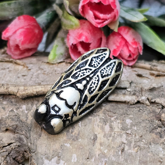 Das Bild zeigt einen handgefertigten Zikaden-Cabochon aus Kunstharz in schwarzer und rosé-silberner Farbgebung. Die Zikade hat eine flache Rückseite mit einem kleinen Loch zwischen den Augen, ideal für die Anbringung einer Anhängeröse.