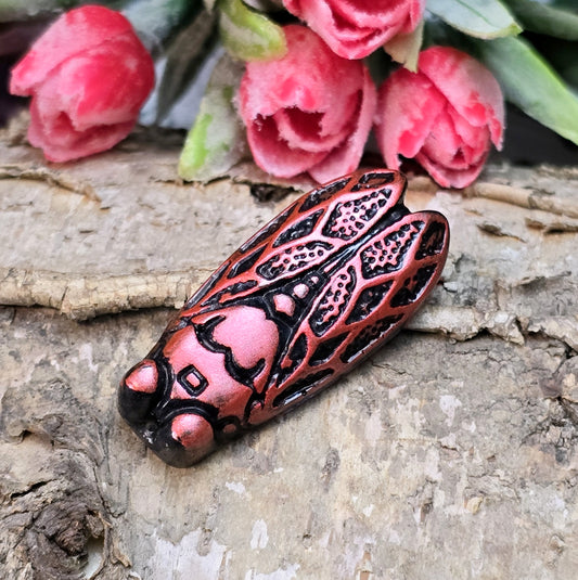 Das Bild zeigt einen handgefertigten Zikaden-Cabochon aus Kunstharz in einer eleganten Karminrot-Optik. Die Zikade hat eine flache Rückseite mit einem kleinen Loch zwischen den Augen, das sich perfekt für die Anbringung einer Anhängeröse eignet.