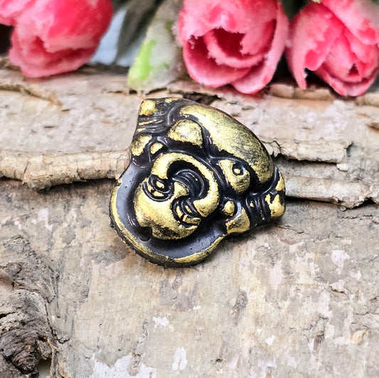 Das Bild zeigt einen handgefertigten Cabochon in Form eines lachenden Buddhas aus Kunstharz. Der Buddha hat eine schwarze Oberfläche mit bronzefarbenen Details und ein kleines Loch am oberen Rand. Die Rückseite ist flach, ideal für die Verwendung als Anhänger oder in Bastelprojekten.