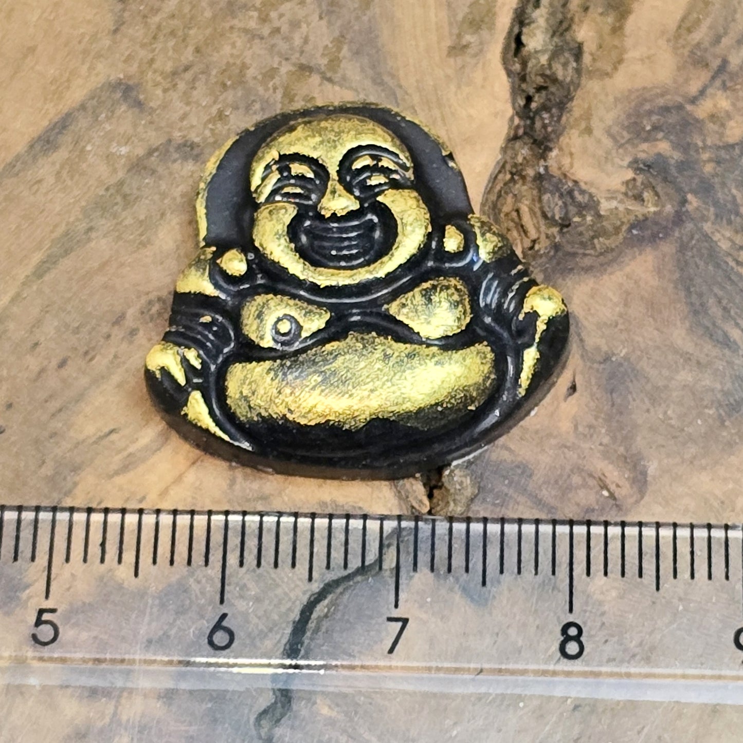 Das Bild zeigt einen handgefertigten Cabochon in Form eines lachenden Buddhas aus Kunstharz. Der Buddha hat eine schwarze Oberfläche mit bronzefarbenen Details und ein kleines Loch am oberen Rand. Die Rückseite ist flach, ideal für die Verwendung als Anhänger oder in Bastelprojekten.