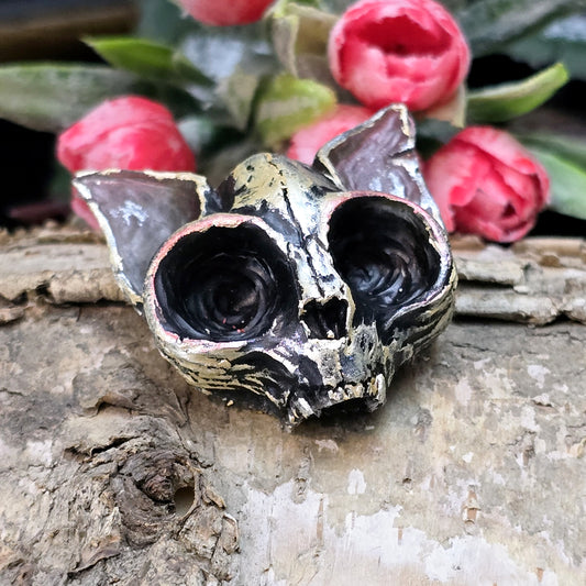 Auf dem Bild ist ein handgefertigter Fledermausschädel aus Kunstharz zu sehen. Der kleine, detailreich gestaltete Schädel ist filigran modelliert und wirkt täuschend echt. Seine Oberfläche ist farblich veredelt, wodurch die feinen Konturen der Knochenstruktur betont werden. Die Rückseite ist flach, sodass er vielseitig verarbeitet werden kann. Der Schädel liegt auf einer dunklen Unterlage, die seinen mystischen Charakter unterstreicht. Er eignet sich perfekt für Schmuckdesign, Bastelprojekte oder als einzig