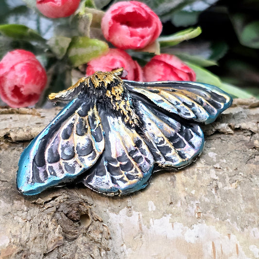 Auf dem Bild ist ein handgefertigter Cabochon in Form einer Deathmoth-Motte zu sehen. Das detailreiche Kunstharzstück zeigt die ausgebreiteten Flügel der mystischen Motte, verziert mit feinen Mustern und Schattierungen. Die Farbgebung betont die Struktur, wodurch das Motiv lebendig wirkt. Die Rückseite ist flach, sodass es sich gut für Schmuck oder Bastelarbeiten eignet. Die Motte liegt auf einem dunklen Untergrund, was ihren geheimnisvollen Charakter unterstreicht.