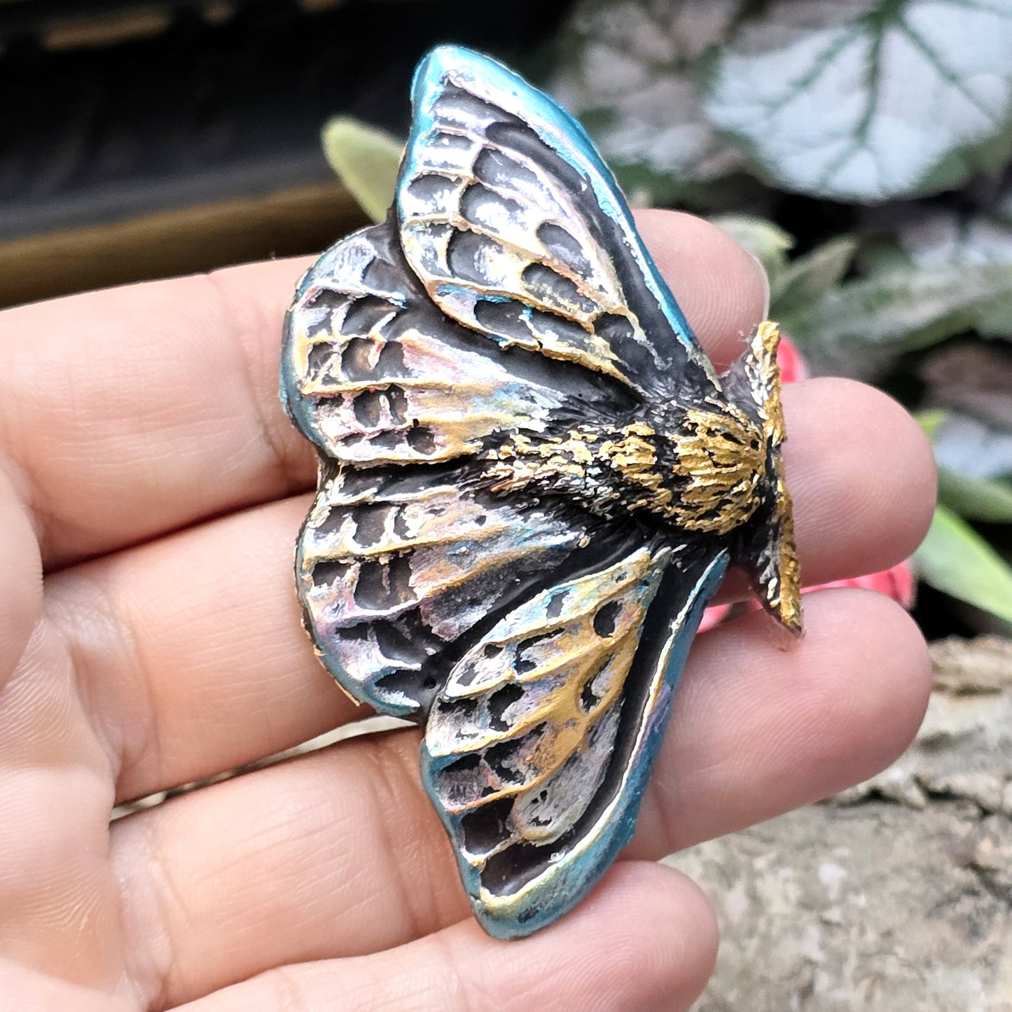 Auf dem Bild ist ein handgefertigter Cabochon in Form einer Deathmoth-Motte zu sehen. Das detailreiche Kunstharzstück zeigt die ausgebreiteten Flügel der mystischen Motte, verziert mit feinen Mustern und Schattierungen. Die Farbgebung betont die Struktur, wodurch das Motiv lebendig wirkt. Die Rückseite ist flach, sodass es sich gut für Schmuck oder Bastelarbeiten eignet. Die Motte liegt auf einem dunklen Untergrund, was ihren geheimnisvollen Charakter unterstreicht.