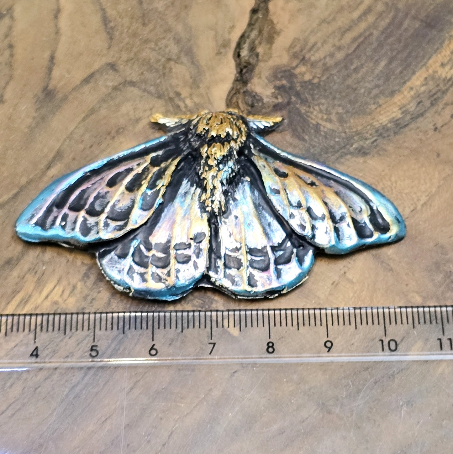 Auf dem Bild ist ein handgefertigter Cabochon in Form einer Deathmoth-Motte zu sehen. Das detailreiche Kunstharzstück zeigt die ausgebreiteten Flügel der mystischen Motte, verziert mit feinen Mustern und Schattierungen. Die Farbgebung betont die Struktur, wodurch das Motiv lebendig wirkt. Die Rückseite ist flach, sodass es sich gut für Schmuck oder Bastelarbeiten eignet. Die Motte liegt auf einem dunklen Untergrund, was ihren geheimnisvollen Charakter unterstreicht.