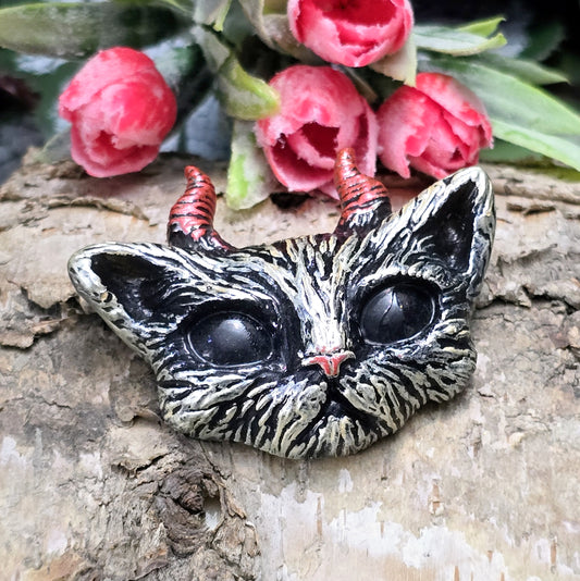 Auf dem Bild ist ein handgefertigter Katzenkopf mit Hörnern aus Kunstharz zu sehen. Der Kopf ist mit handbemalten Details verziert und hat eine flache Rückseite, die ideal für Schmuckherstellung oder Bastelprojekte geeignet ist.