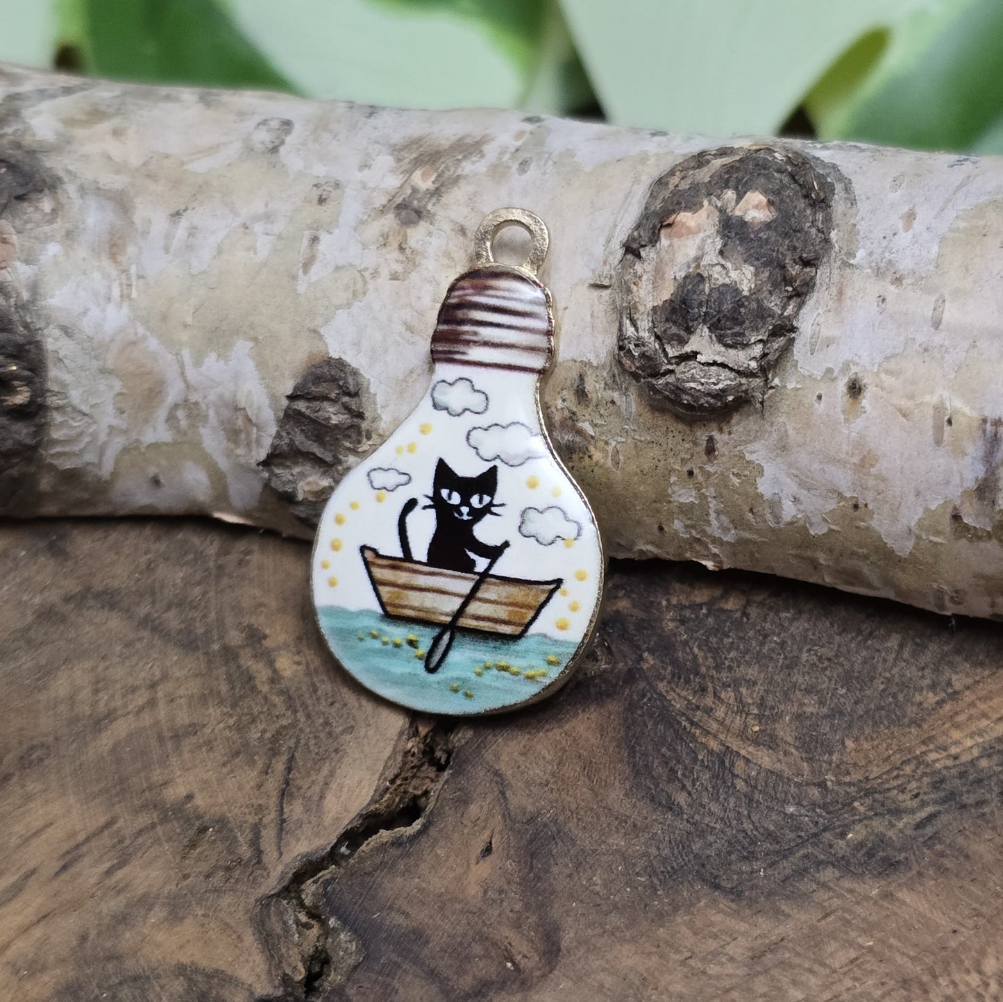 Das Bild zeigt einen goldfarbenen Anhänger in Glühbirnenform, auf dem ein Katzenmotiv abgebildet ist. Eine Katze sitzt in einem Ruderboot und fährt auf einem malerischen See. Das filigrane Design ist von einer goldenen Umrandung eingefasst, die dem Anhänger eine edle Note verleiht. Der Anhänger hat eine Größe von 27mm und eignet sich hervorragend für Schmuckherstellung oder DIY-Projekte.