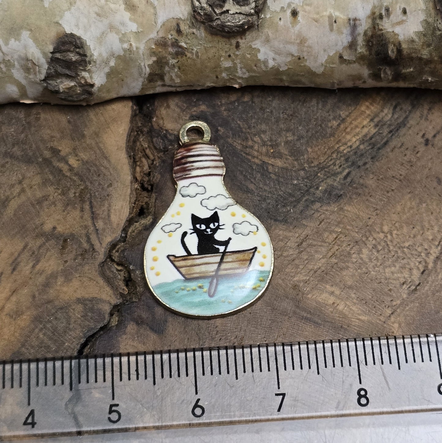 Das Bild zeigt einen goldfarbenen Anhänger in Glühbirnenform, auf dem ein Katzenmotiv abgebildet ist. Eine Katze sitzt in einem Ruderboot und fährt auf einem malerischen See. Das filigrane Design ist von einer goldenen Umrandung eingefasst, die dem Anhänger eine edle Note verleiht. Der Anhänger hat eine Größe von 27mm und eignet sich hervorragend für Schmuckherstellung oder DIY-Projekte.