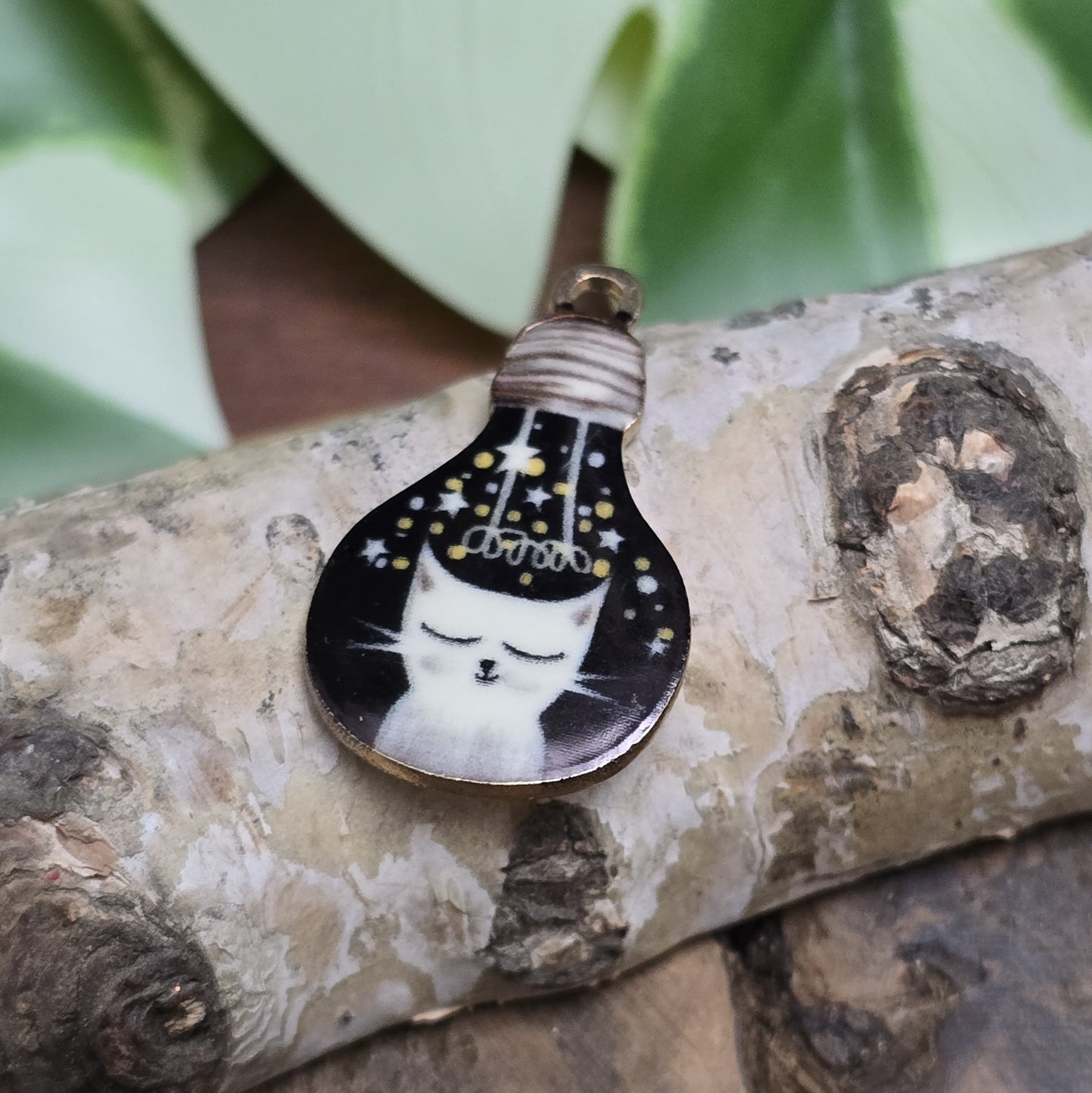 Das Bild zeigt einen goldfarbenen Anhänger in Glühbirnenform, auf dem eine weiße Katze mit geschlossenen Augen abgebildet ist. Die Katze ist nur zur Hälfte sichtbar und sitzt auf einem schwarzen Hintergrund, der die Eleganz des Designs unterstreicht. Der Anhänger hat eine Größe von 27mm und ist von einer goldenen Umrandung eingefasst, die ihm eine edle Ausstrahlung verleiht. Dieser Anhänger eignet sich hervorragend für Schmuckherstellung und kreative DIY-Projekte.