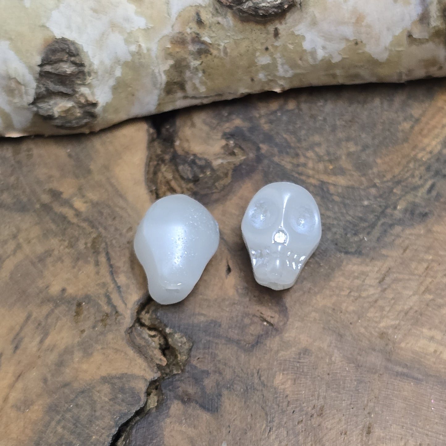 Das Bild zeigt zwei matt gräulich weiße Glasperlen in Totenkopf-Form. Die Perlen sind dreidimensional gestaltet, sodass die Schädelstruktur mit Augenhöhlen, Nase und Kiefer erkennbar ist. Die Bohrung verläuft von oben nach unten, sodass sie ideal für Schmuckprojekte verwendet werden können. Durch die matte Oberfläche wirkt das Design besonders stilvoll und modern.
