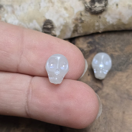 Das Bild zeigt zwei matt gräulich weiße Glasperlen in Totenkopf-Form. Die Perlen sind dreidimensional gestaltet, sodass die Schädelstruktur mit Augenhöhlen, Nase und Kiefer erkennbar ist. Die Bohrung verläuft von oben nach unten, sodass sie ideal für Schmuckprojekte verwendet werden können. Durch die matte Oberfläche wirkt das Design besonders stilvoll und modern.