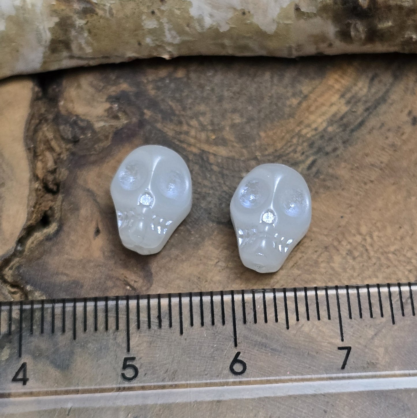 Das Bild zeigt zwei matt gräulich weiße Glasperlen in Totenkopf-Form. Die Perlen sind dreidimensional gestaltet, sodass die Schädelstruktur mit Augenhöhlen, Nase und Kiefer erkennbar ist. Die Bohrung verläuft von oben nach unten, sodass sie ideal für Schmuckprojekte verwendet werden können. Durch die matte Oberfläche wirkt das Design besonders stilvoll und modern.