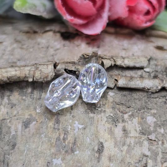Das Bild zeigt zwei schimmernde Glasperlen in Totenkopf-Form mit Crystal-AB-Beschichtung. Die Perlen sind dreidimensional gestaltet, sodass Details wie Augenhöhlen, Nase und Kiefer deutlich erkennbar sind. Ihre Oberfläche reflektiert das Licht in irisierenden Farben, die je nach Blickwinkel changieren. Die Bohrung verläuft von oben nach unten, sodass sie ideal für Schmuckprojekte geeignet sind.