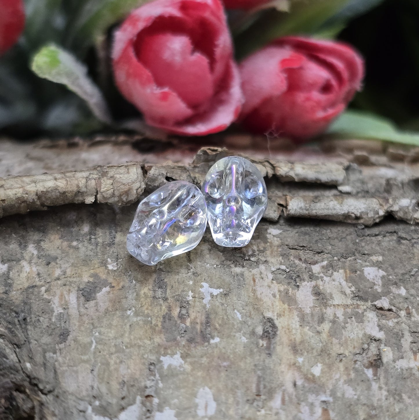 Das Bild zeigt zwei schimmernde Glasperlen in Totenkopf-Form mit Crystal-AB-Beschichtung. Die Perlen sind dreidimensional gestaltet, sodass Details wie Augenhöhlen, Nase und Kiefer deutlich erkennbar sind. Ihre Oberfläche reflektiert das Licht in irisierenden Farben, die je nach Blickwinkel changieren. Die Bohrung verläuft von oben nach unten, sodass sie ideal für Schmuckprojekte geeignet sind.