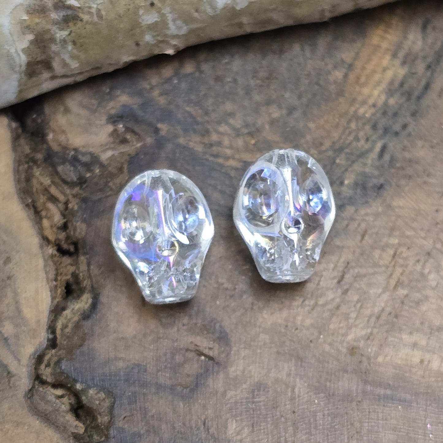 Das Bild zeigt zwei schimmernde Glasperlen in Totenkopf-Form mit Crystal-AB-Beschichtung. Die Perlen sind dreidimensional gestaltet, sodass Details wie Augenhöhlen, Nase und Kiefer deutlich erkennbar sind. Ihre Oberfläche reflektiert das Licht in irisierenden Farben, die je nach Blickwinkel changieren. Die Bohrung verläuft von oben nach unten, sodass sie ideal für Schmuckprojekte geeignet sind.