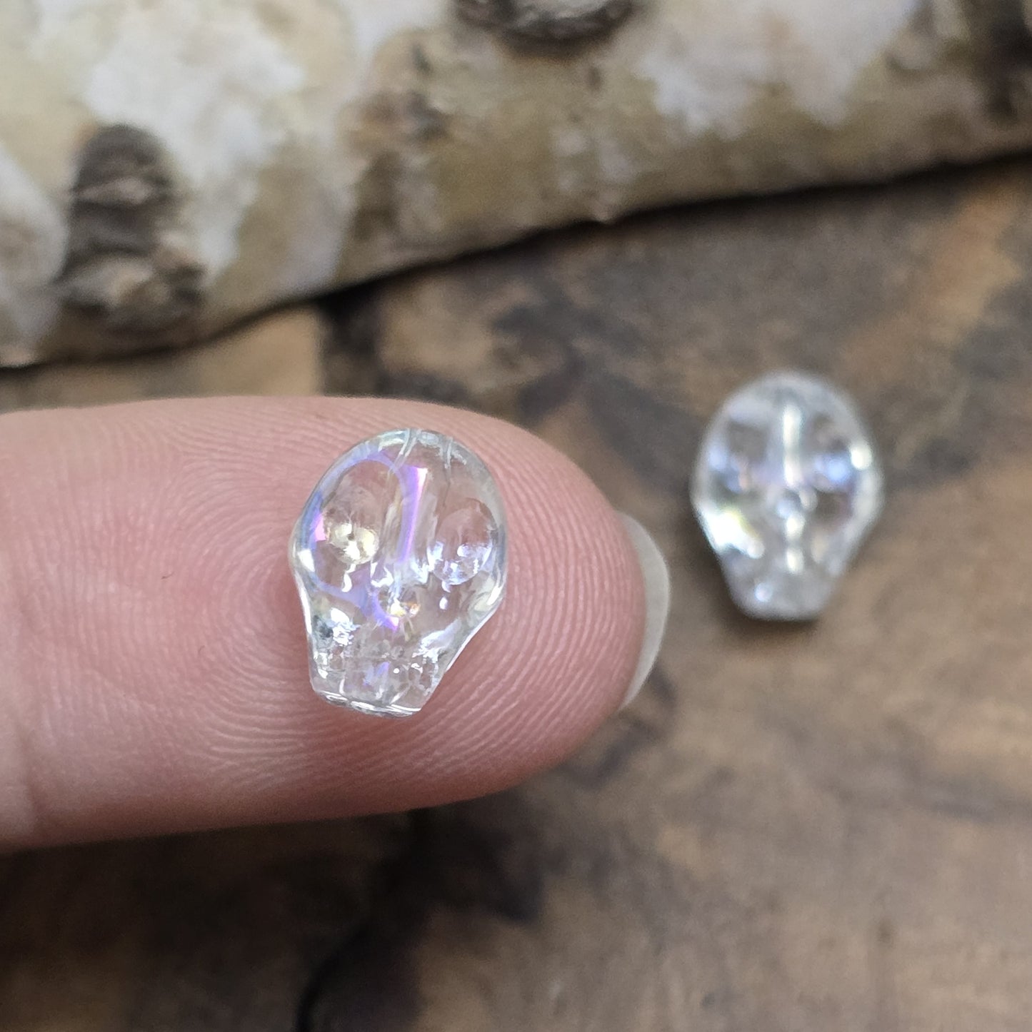 Das Bild zeigt zwei schimmernde Glasperlen in Totenkopf-Form mit Crystal-AB-Beschichtung. Die Perlen sind dreidimensional gestaltet, sodass Details wie Augenhöhlen, Nase und Kiefer deutlich erkennbar sind. Ihre Oberfläche reflektiert das Licht in irisierenden Farben, die je nach Blickwinkel changieren. Die Bohrung verläuft von oben nach unten, sodass sie ideal für Schmuckprojekte geeignet sind.