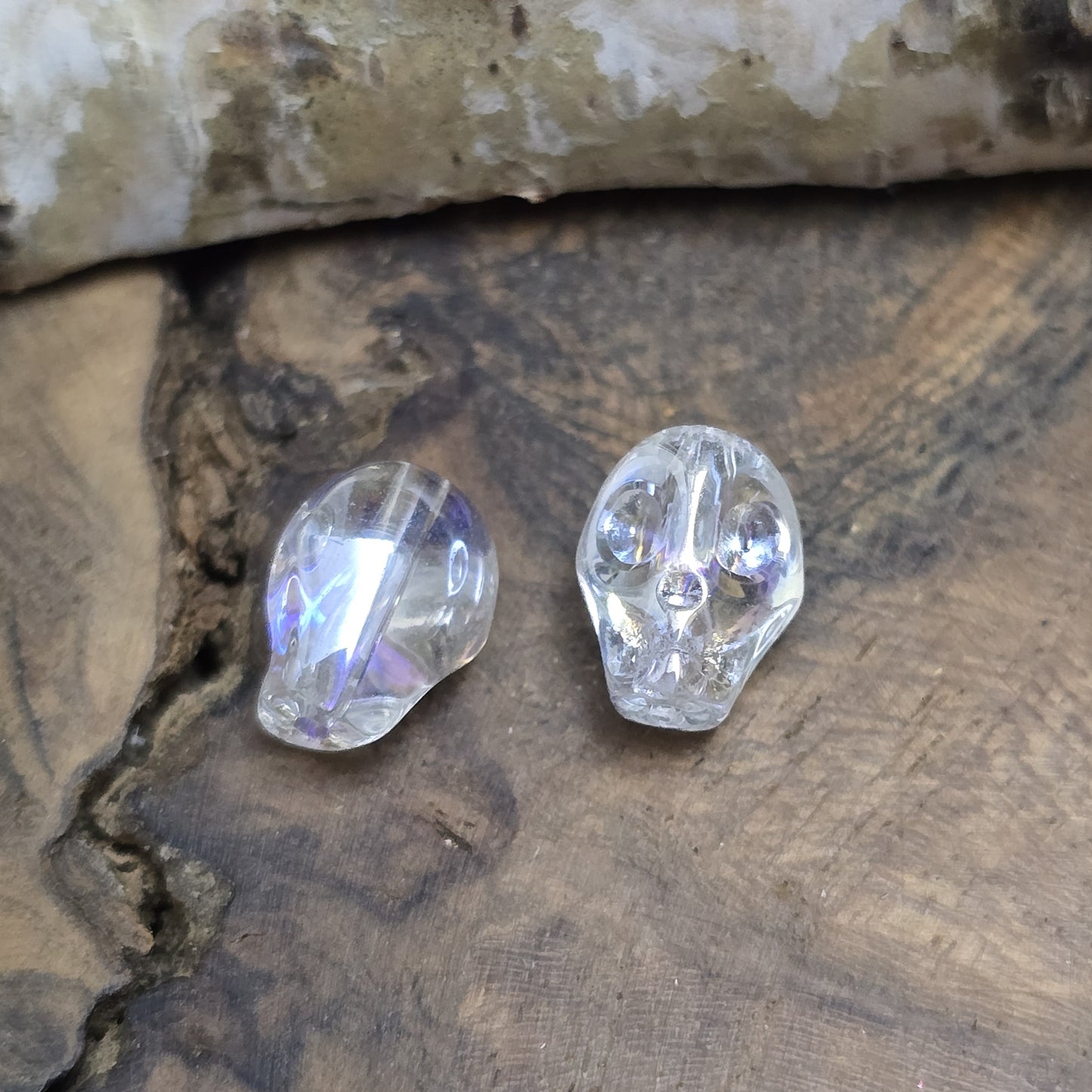 Das Bild zeigt zwei schimmernde Glasperlen in Totenkopf-Form mit Crystal-AB-Beschichtung. Die Perlen sind dreidimensional gestaltet, sodass Details wie Augenhöhlen, Nase und Kiefer deutlich erkennbar sind. Ihre Oberfläche reflektiert das Licht in irisierenden Farben, die je nach Blickwinkel changieren. Die Bohrung verläuft von oben nach unten, sodass sie ideal für Schmuckprojekte geeignet sind.