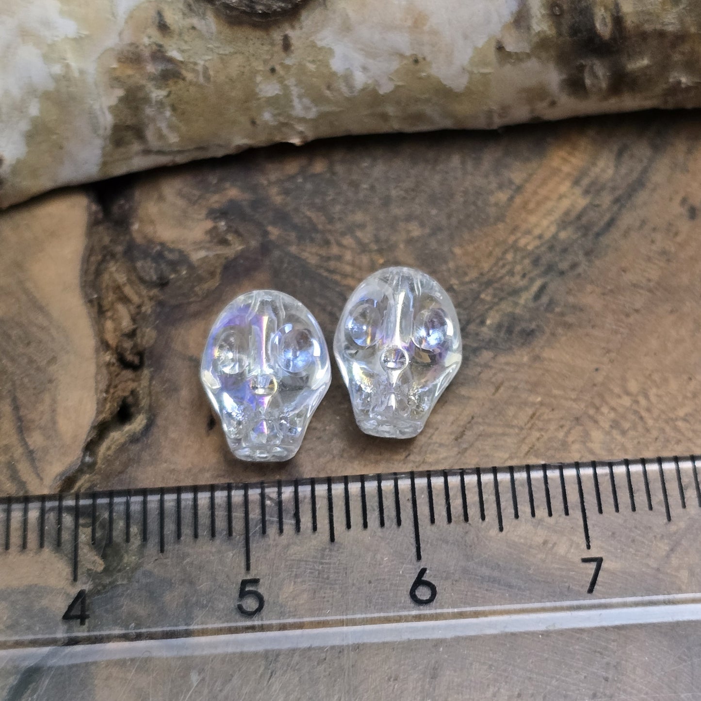 Das Bild zeigt zwei schimmernde Glasperlen in Totenkopf-Form mit Crystal-AB-Beschichtung. Die Perlen sind dreidimensional gestaltet, sodass Details wie Augenhöhlen, Nase und Kiefer deutlich erkennbar sind. Ihre Oberfläche reflektiert das Licht in irisierenden Farben, die je nach Blickwinkel changieren. Die Bohrung verläuft von oben nach unten, sodass sie ideal für Schmuckprojekte geeignet sind.
