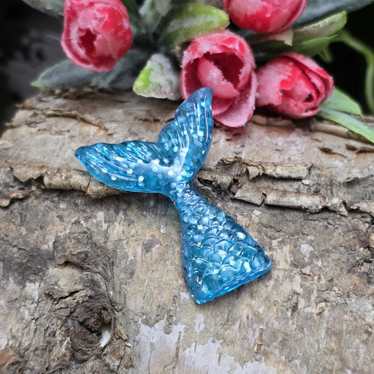 Das Bild zeigt einen handgefertigten Cabochon in Form einer Meerjungfrauenschwanzflosse. Der Anhänger besteht aus Kunstharz und leuchtet in einem schimmernden Aqua Blau mit feinem Glitter. Die geschwungene Form und die detailreiche Struktur verleihen ihm eine elegante, maritime Optik.