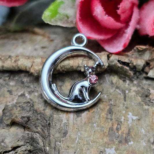 Das Bild zeigt einen Anhänger in Silberfarbe, der eine Katze im Mond darstellt. Die Katze ist elegant im Mond sitzend, begleitet von einem funkelnden rosé Strass-Stein. Der Anhänger ist ca. 20mm groß und eignet sich perfekt für die Schmuckherstellung oder als kreatives Accessoire.