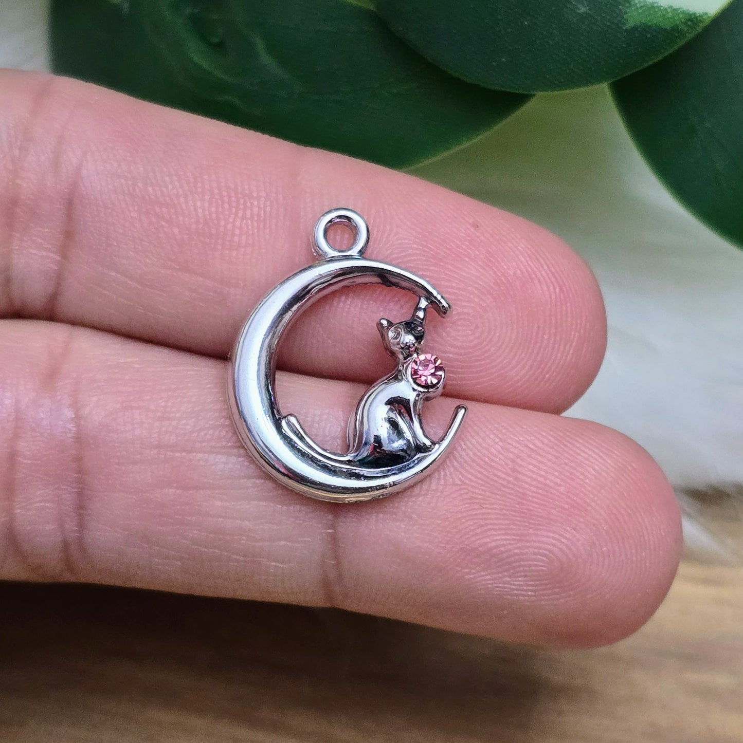 Das Bild zeigt einen Anhänger in Silberfarbe, der eine Katze im Mond darstellt. Die Katze ist elegant im Mond sitzend, begleitet von einem funkelnden rosé Strass-Stein. Der Anhänger ist ca. 20mm groß und eignet sich perfekt für die Schmuckherstellung oder als kreatives Accessoire.