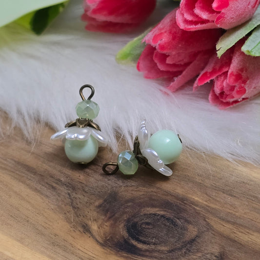 Das Bild zeigt zwei handgefertigte Anhänger mit lindgrünen Glasperlen. Jede Perle ist von einem perlweißen Blütenkäppchen aus Acryl umgeben, während eine facettierte Kristallglasperle in Lindgrün für funkelnde Akzente sorgt. Eine antik bronzefarbene Perlenkappe verleiht dem Design einen nostalgischen Charme. Die Anhänger haben eine Größe von ca. 20mm und eignen sich ideal für Schmuckkreationen im Vintage- oder romantischen Stil.