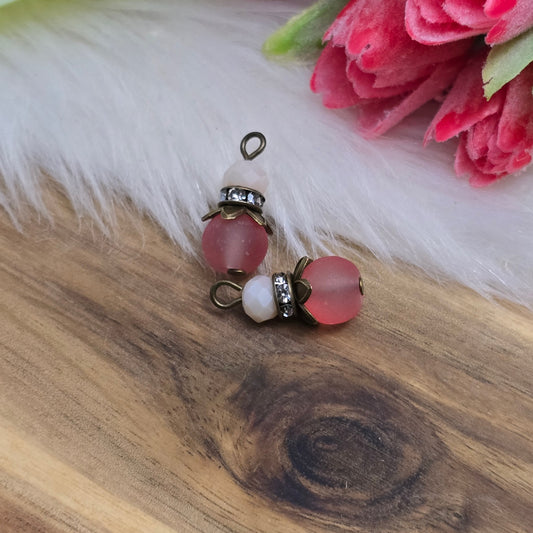 Das Bild zeigt zwei handgefertigte Anhänger mit einer matten, transparenten lachsfarbenen Glasperle als Hauptbestandteil. Darüber sitzt eine kleinere, facettierte rosa Kristallglasperle, die sanft im Licht schimmert. Ein glänzendes Strassrondell trennt die beiden Perlen und verleiht dem Design einen funkelnden Akzent. Abgerundet wird der Anhänger durch eine antik bronzefarbene Perlenkappe, die dem Schmuckstück einen edlen Vintage-Charakter verleiht. Die Anhänger haben eine Größe von ca. 18mm und eignen sich