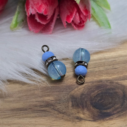Das Bild zeigt zwei handgefertigte Anhänger mit einer Kombination aus transparenten hellblauen Glasperlen und opaken hellblauen Kristallglasperlen. Ein funkelndes Strassrondell setzt glitzernde Akzente, während antik bronzefarbige Metallelemente dem Design einen charmanten Vintage-Touch verleihen. Die filigranen Details und die harmonische Farbzusammenstellung machen die Anhänger zu einem stilvollen Blickfang für Schmuckkreationen.