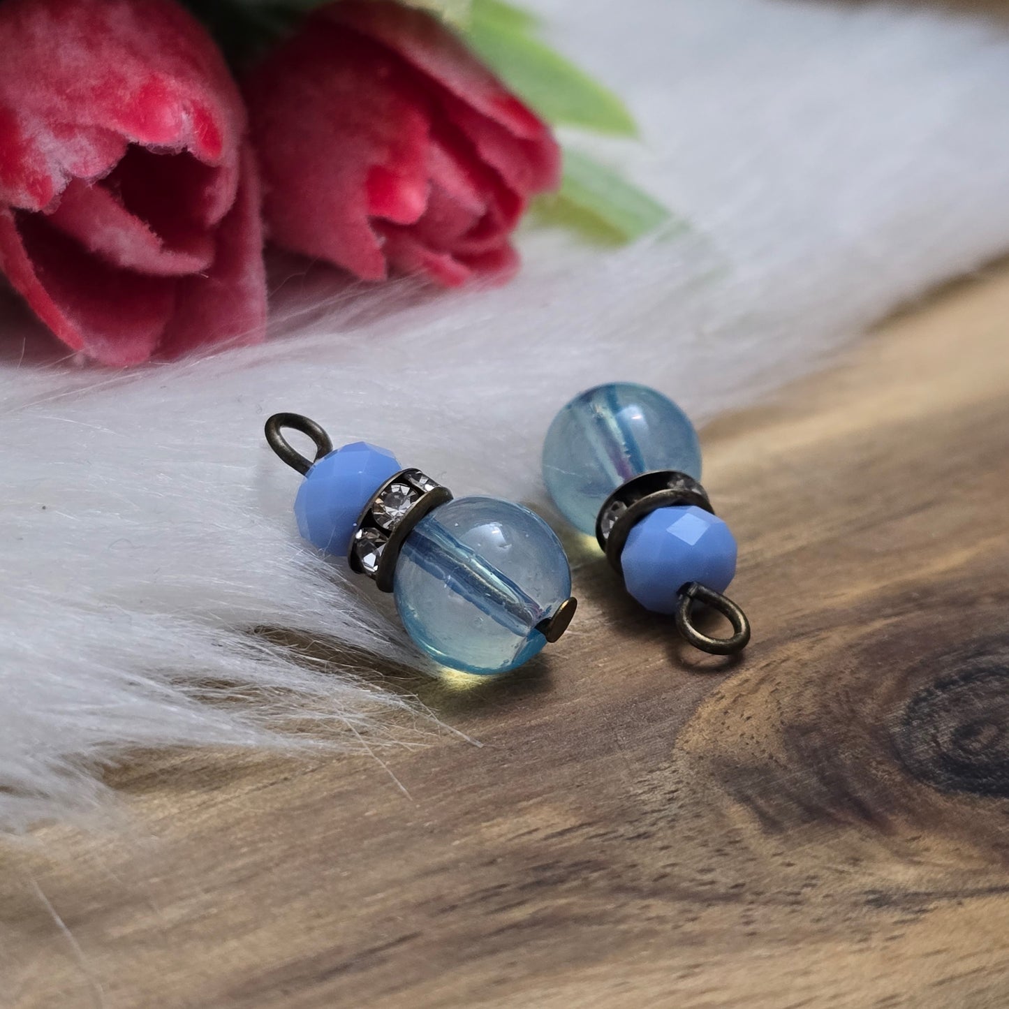 Das Bild zeigt zwei handgefertigte Anhänger mit einer Kombination aus transparenten hellblauen Glasperlen und opaken hellblauen Kristallglasperlen. Ein funkelndes Strassrondell setzt glitzernde Akzente, während antik bronzefarbige Metallelemente dem Design einen charmanten Vintage-Touch verleihen. Die filigranen Details und die harmonische Farbzusammenstellung machen die Anhänger zu einem stilvollen Blickfang für Schmuckkreationen.