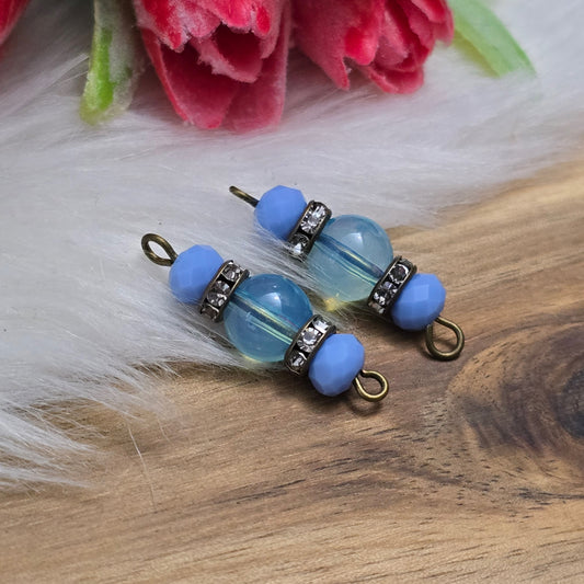Das Bild zeigt zwei handgefertigte Verbinder mit hellblauen Glasperlen. Die Kombination aus einer transparenten hellblauen Glasperle (8mm) und einer opaken hellblauen Kristallglasperle wird durch ein funkelndes Strassrondell ergänzt. Antik bronzefarbene Metallelemente verleihen den Verbindern einen eleganten Vintage-Look. Mit einer Länge von 27mm sind sie ideal für die Schmuckherstellung.
