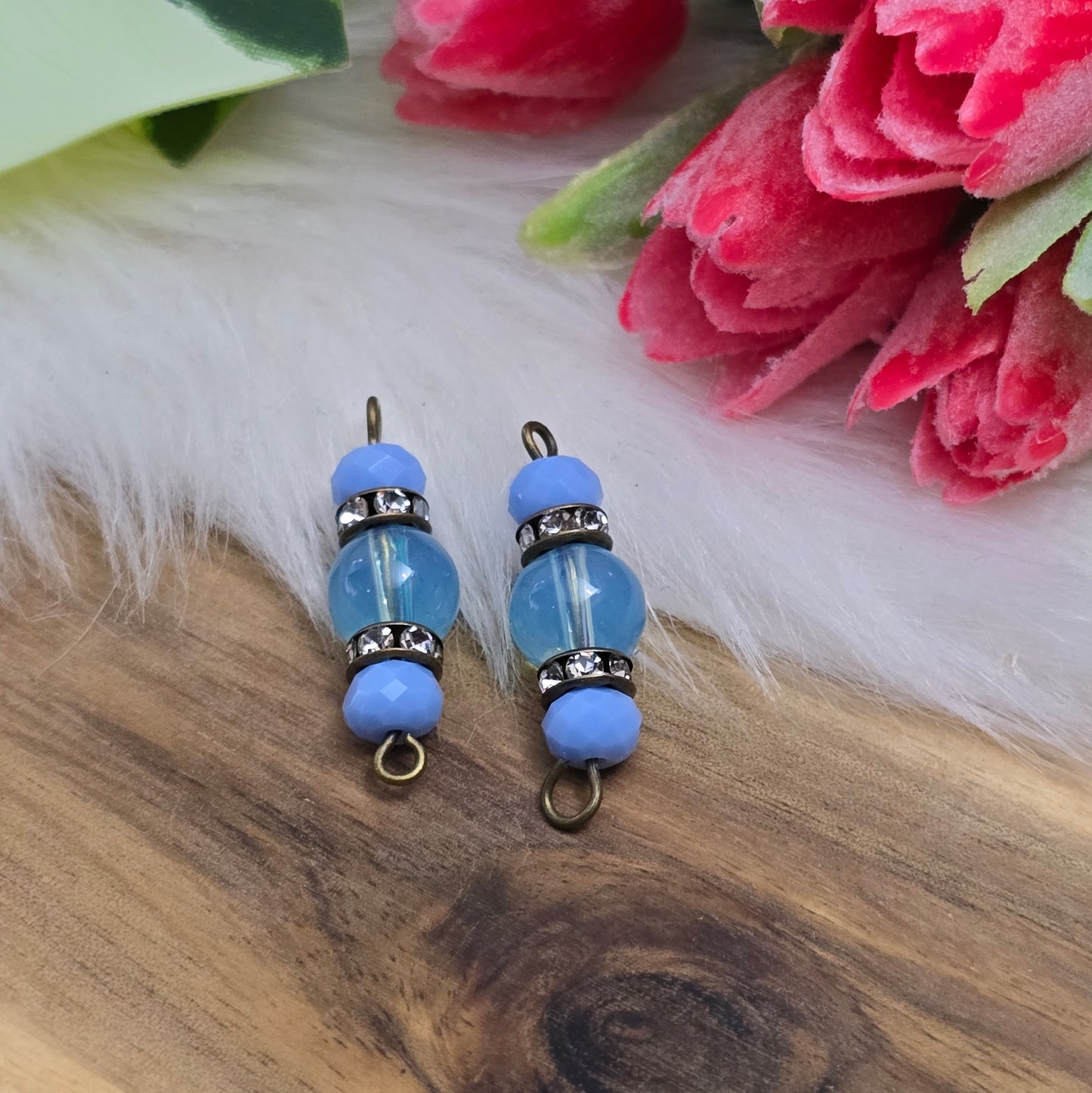 Das Bild zeigt zwei handgefertigte Verbinder mit hellblauen Glasperlen. Die Kombination aus einer transparenten hellblauen Glasperle (8mm) und einer opaken hellblauen Kristallglasperle wird durch ein funkelndes Strassrondell ergänzt. Antik bronzefarbene Metallelemente verleihen den Verbindern einen eleganten Vintage-Look. Mit einer Länge von 27mm sind sie ideal für die Schmuckherstellung.