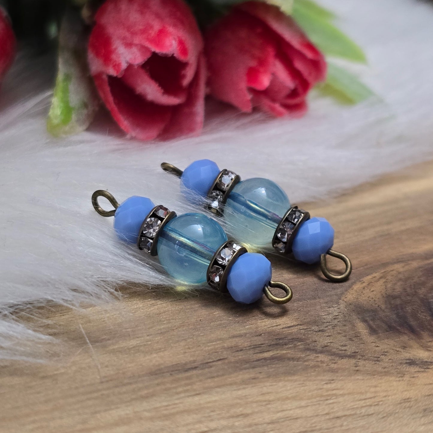 Das Bild zeigt zwei handgefertigte Verbinder mit hellblauen Glasperlen. Die Kombination aus einer transparenten hellblauen Glasperle (8mm) und einer opaken hellblauen Kristallglasperle wird durch ein funkelndes Strassrondell ergänzt. Antik bronzefarbene Metallelemente verleihen den Verbindern einen eleganten Vintage-Look. Mit einer Länge von 27mm sind sie ideal für die Schmuckherstellung.
