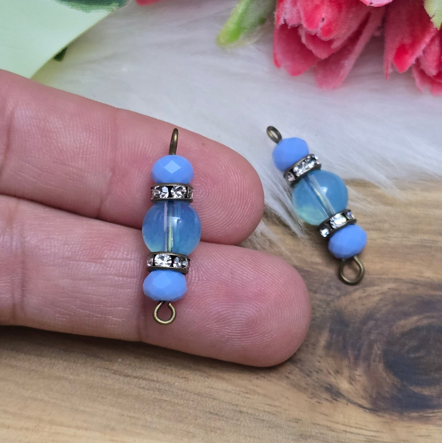 Das Bild zeigt zwei handgefertigte Verbinder mit hellblauen Glasperlen. Die Kombination aus einer transparenten hellblauen Glasperle (8mm) und einer opaken hellblauen Kristallglasperle wird durch ein funkelndes Strassrondell ergänzt. Antik bronzefarbene Metallelemente verleihen den Verbindern einen eleganten Vintage-Look. Mit einer Länge von 27mm sind sie ideal für die Schmuckherstellung.