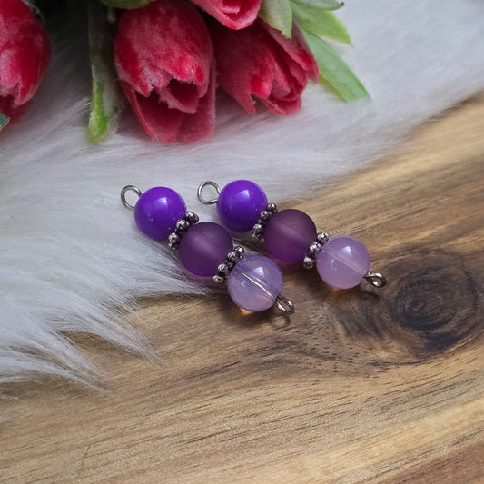 Das Bild zeigt zwei handgefertigte Verbinder mit lila und violetten Glasperlen in unterschiedlichen Ausführungen – eine opake, eine matte und eine milchig transparente. Die Perlen sind harmonisch angeordnet und werden von detailreich verzierten, antik-silberfarbenen Metallelementen eingefasst. Die filigranen Zwischenelemente verleihen den Verbindern eine edle Vintage-Optik. Mit einer Länge von ca. 33mm eignen sie sich perfekt für kreative Schmuckdesigns wie Armbänder, Ketten oder Ohrringe.