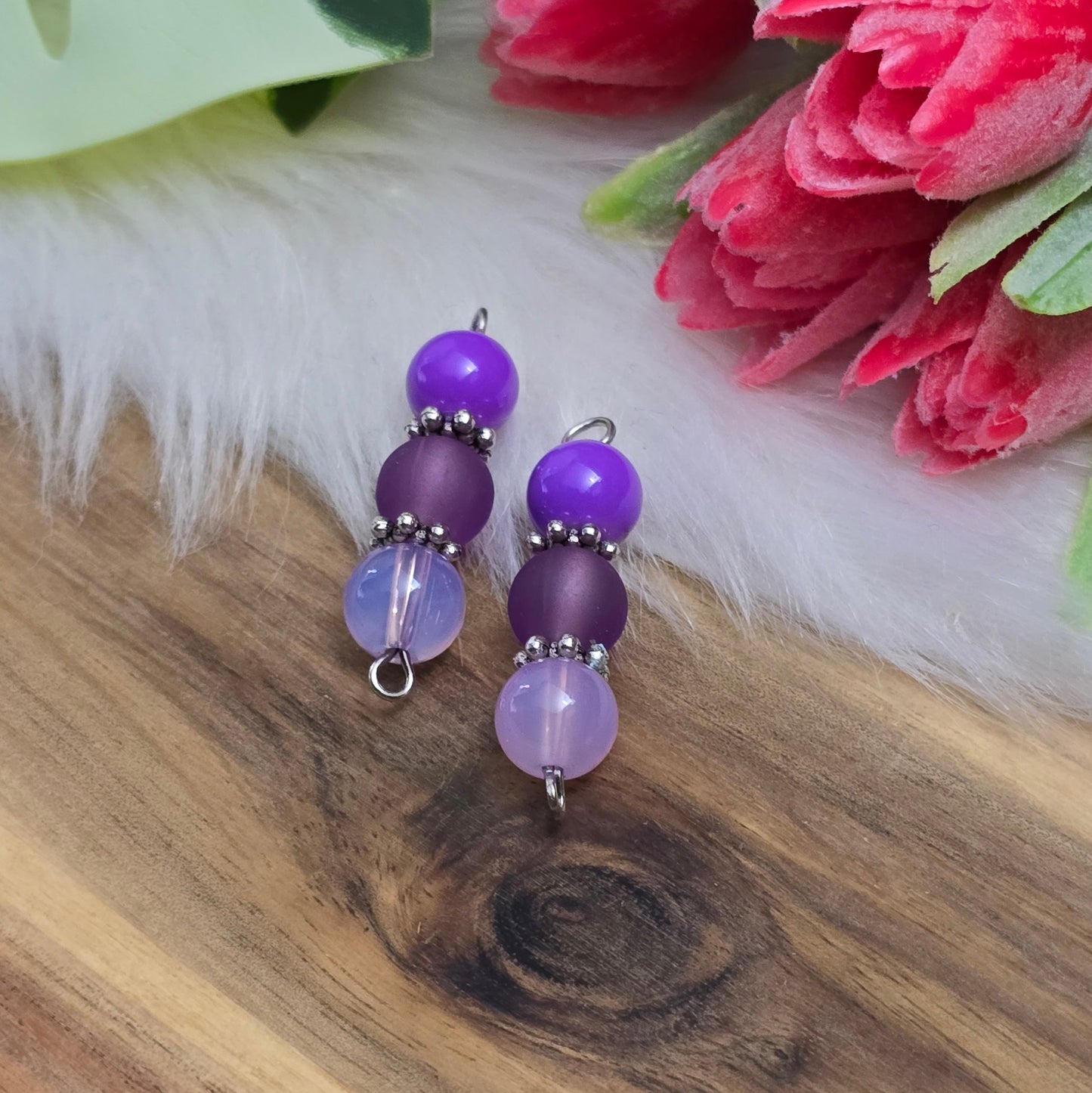 Das Bild zeigt zwei handgefertigte Verbinder mit lila und violetten Glasperlen in unterschiedlichen Ausführungen – eine opake, eine matte und eine milchig transparente. Die Perlen sind harmonisch angeordnet und werden von detailreich verzierten, antik-silberfarbenen Metallelementen eingefasst. Die filigranen Zwischenelemente verleihen den Verbindern eine edle Vintage-Optik. Mit einer Länge von ca. 33mm eignen sie sich perfekt für kreative Schmuckdesigns wie Armbänder, Ketten oder Ohrringe.
