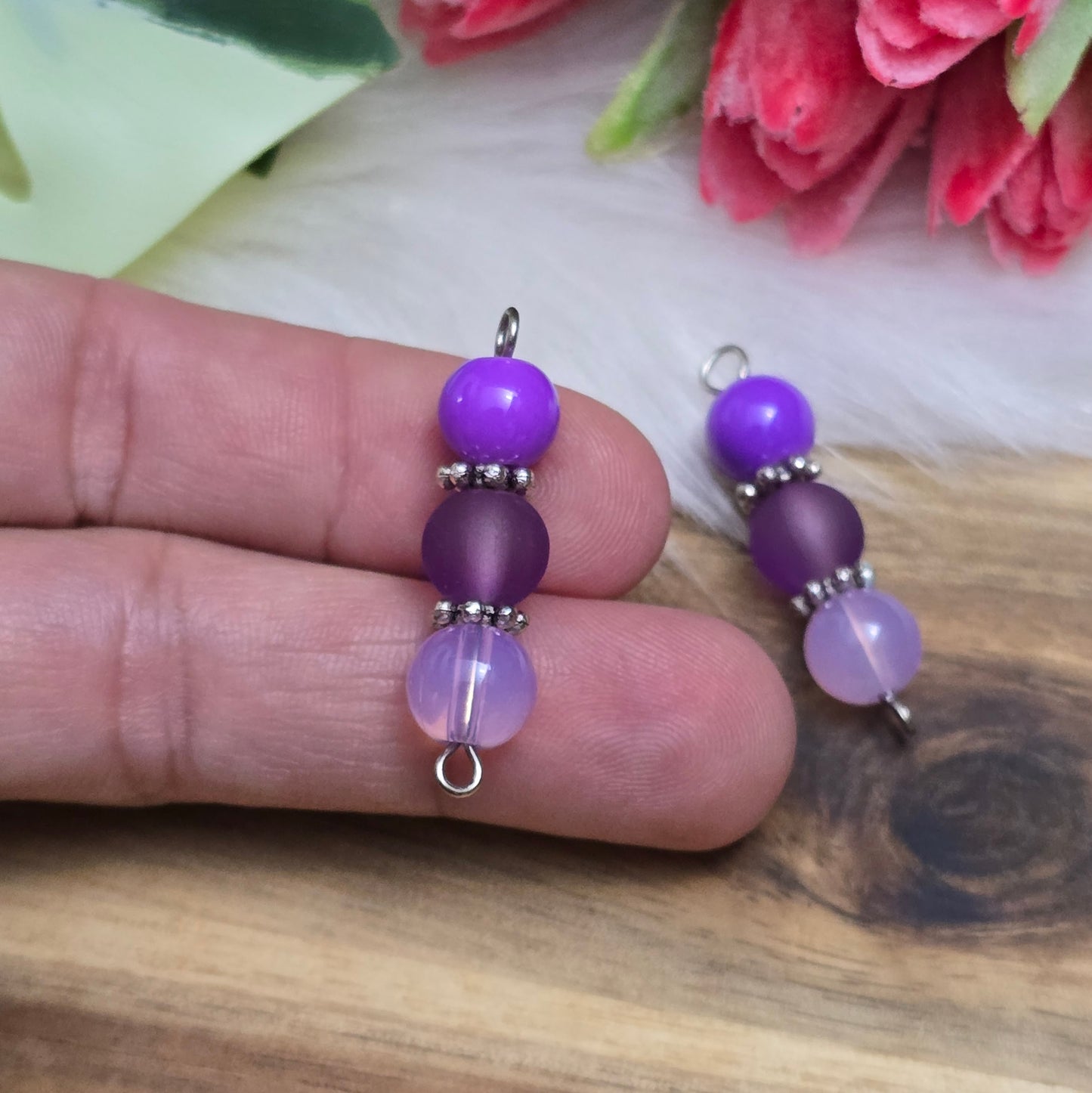 Das Bild zeigt zwei handgefertigte Verbinder mit lila und violetten Glasperlen in unterschiedlichen Ausführungen – eine opake, eine matte und eine milchig transparente. Die Perlen sind harmonisch angeordnet und werden von detailreich verzierten, antik-silberfarbenen Metallelementen eingefasst. Die filigranen Zwischenelemente verleihen den Verbindern eine edle Vintage-Optik. Mit einer Länge von ca. 33mm eignen sie sich perfekt für kreative Schmuckdesigns wie Armbänder, Ketten oder Ohrringe.