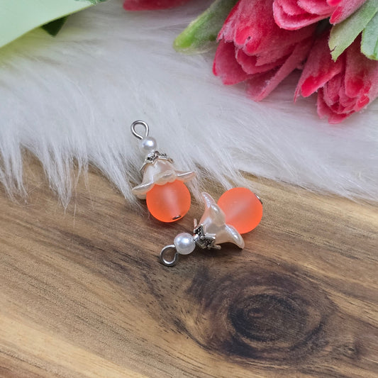 Das Bild zeigt zwei handgemachte Anhänger mit matten, orangefarbenen Glasperlen. Jedes Stück verfügt über ein elegantes, weißes Acrylblütenkäppchen, das einen verspielten Akzent setzt. Die antik silberfarbigen Perlenkappen runden das Design harmonisch ab. Mit einem Durchmesser von etwa 20mm sind die Anhänger filigran und ideal für Schmuckprojekte. Die Kombination aus lebendigen Farben und stilvollen Materialien verleiht den Anhängern einen attraktiven Look, der in jeder Schmuckkollektion hervorsticht.