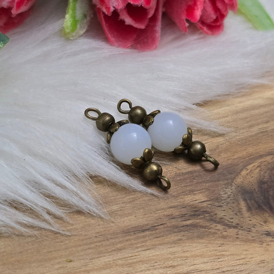 Das Bild zeigt zwei handgefertigte Schmuckverbinder mit milchig weißen Glasperlen und antik bronzefarbenen Metallelementen. Die Perlen haben eine sanft schimmernde Oberfläche, die ihnen eine elegante, edle Optik verleiht. Die filigranen Metallverzierungen sorgen für einen charmanten Vintage-Stil. Die Verbinder sind kompakt und vielseitig einsetzbar – ideal für DIY-Schmuckprojekte wie Halsketten, Armbänder oder Ohrringe. Ein stilvoller Blickfang für kreative Designs! ✨