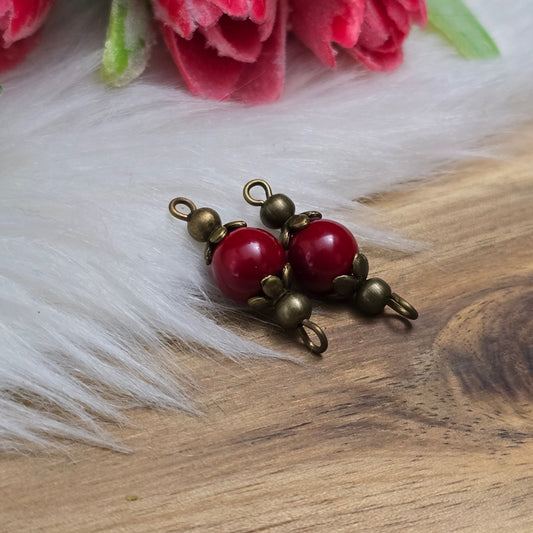 Das Bild zeigt zwei handgefertigte Schmuckverbinder mit tief dunkelroten Glasperlen, die sanft schimmern. Die Perlen sind in detailreiche, antik bronzefarbene Metallelemente eingefasst, die ihnen einen edlen Vintage-Look verleihen. Die filigrane Verarbeitung betont die elegante Form der Verbinder, die sich perfekt für Schmuckdesigns eignen. Die warme Farbkombination aus Dunkelrot und Antik-Bronze sorgt für einen stilvollen, zeitlosen Charme. ✨
