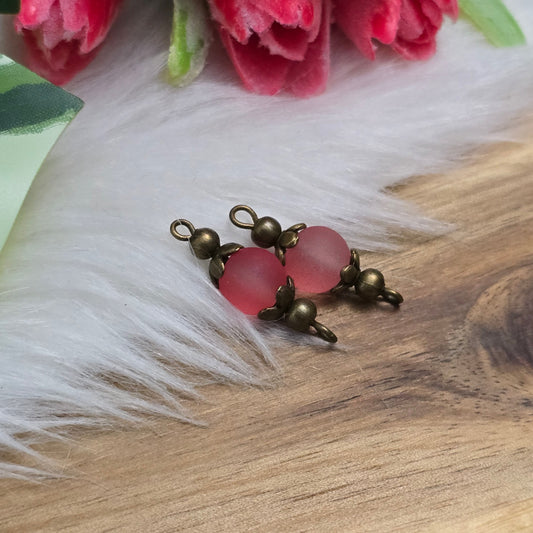 Die Aufnahme zeigt zwei filigrane, handgefertigte Schmuckverbinder mit matten lachsfarbenen Glasperlen. Die sanft schimmernden Perlen sind in kunstvoll verzierten, antik bronzefarbenen Metallelementen eingefasst, die ihnen eine elegante Vintage-Optik verleihen. Die warmen Farbtöne der Glasperlen harmonieren perfekt mit dem dunklen Metall und machen die Verbinder zu einem stilvollen Highlight für Schmuckkreationen.