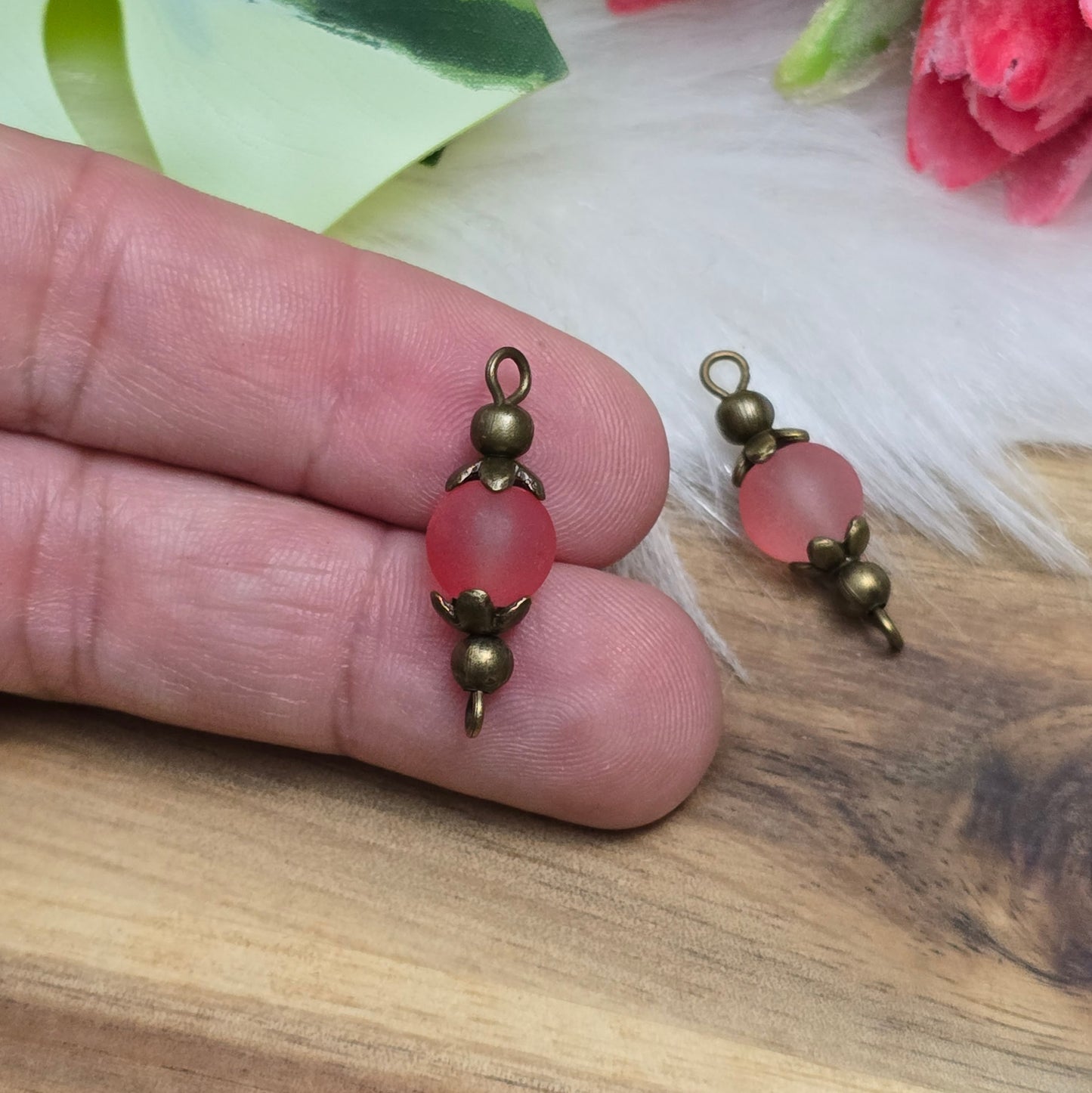 Die Aufnahme zeigt zwei filigrane, handgefertigte Schmuckverbinder mit matten lachsfarbenen Glasperlen. Die sanft schimmernden Perlen sind in kunstvoll verzierten, antik bronzefarbenen Metallelementen eingefasst, die ihnen eine elegante Vintage-Optik verleihen. Die warmen Farbtöne der Glasperlen harmonieren perfekt mit dem dunklen Metall und machen die Verbinder zu einem stilvollen Highlight für Schmuckkreationen.