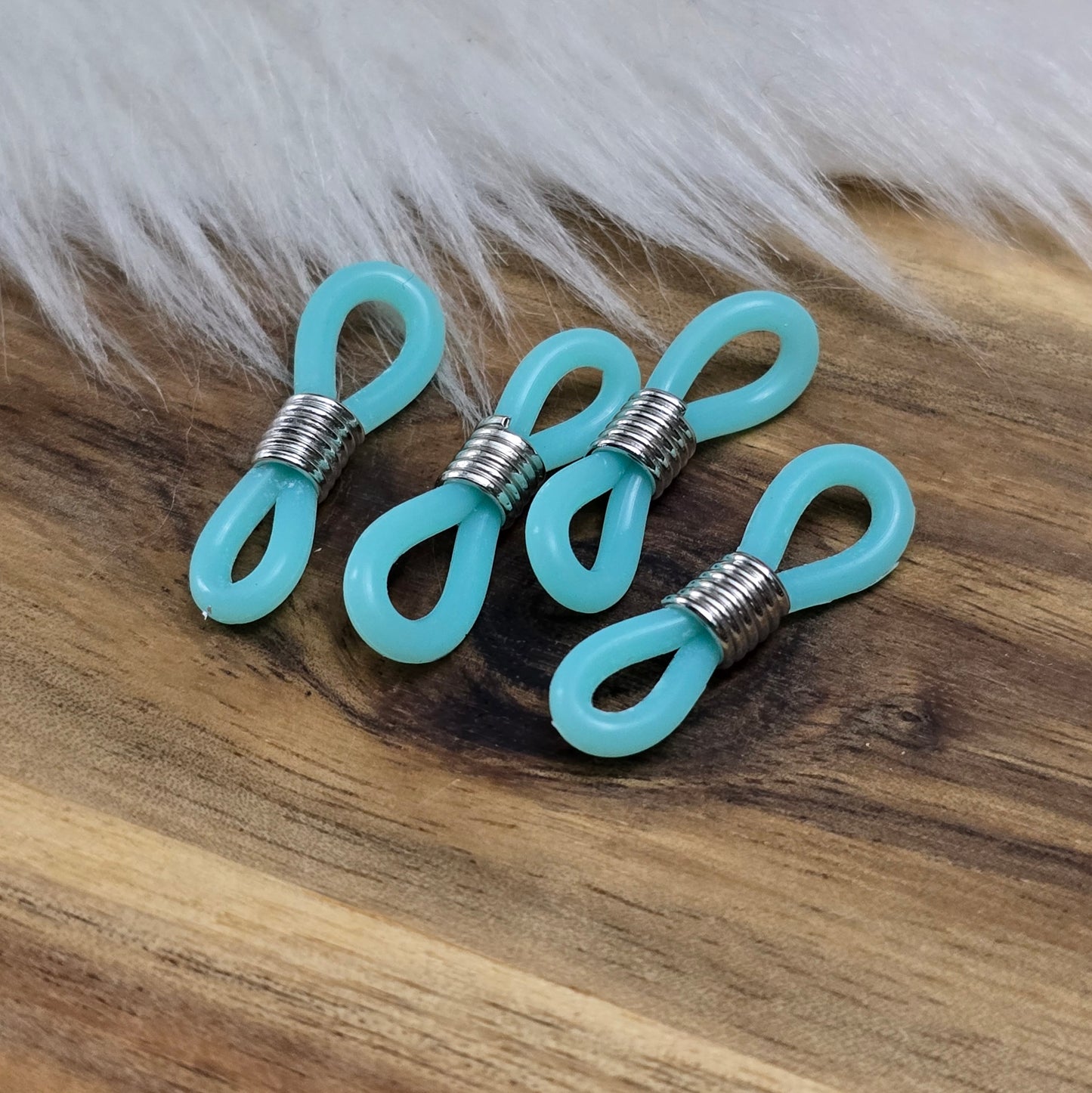 Das Bild zeigt eine Auswahl an Silikonschlaufen für Brillenketten und Maskenhalter in drei Farben: Mint, Rosa und Schwarz. Jede Schlaufe hat eine flexible, weiche Textur und ist so gestaltet, dass sie einfach auf den Bügel der Brille geschoben werden kann. An einem Ende der Schlaufe befindet sich ein kleiner Metallring, der fest angebracht ist, um ein Abrutschen zu verhindern. Die anderen Enden der Schlaufen sind offen, sodass sie mit einem Karabiner oder Biegering leicht an einer Kette befestigt werden kön