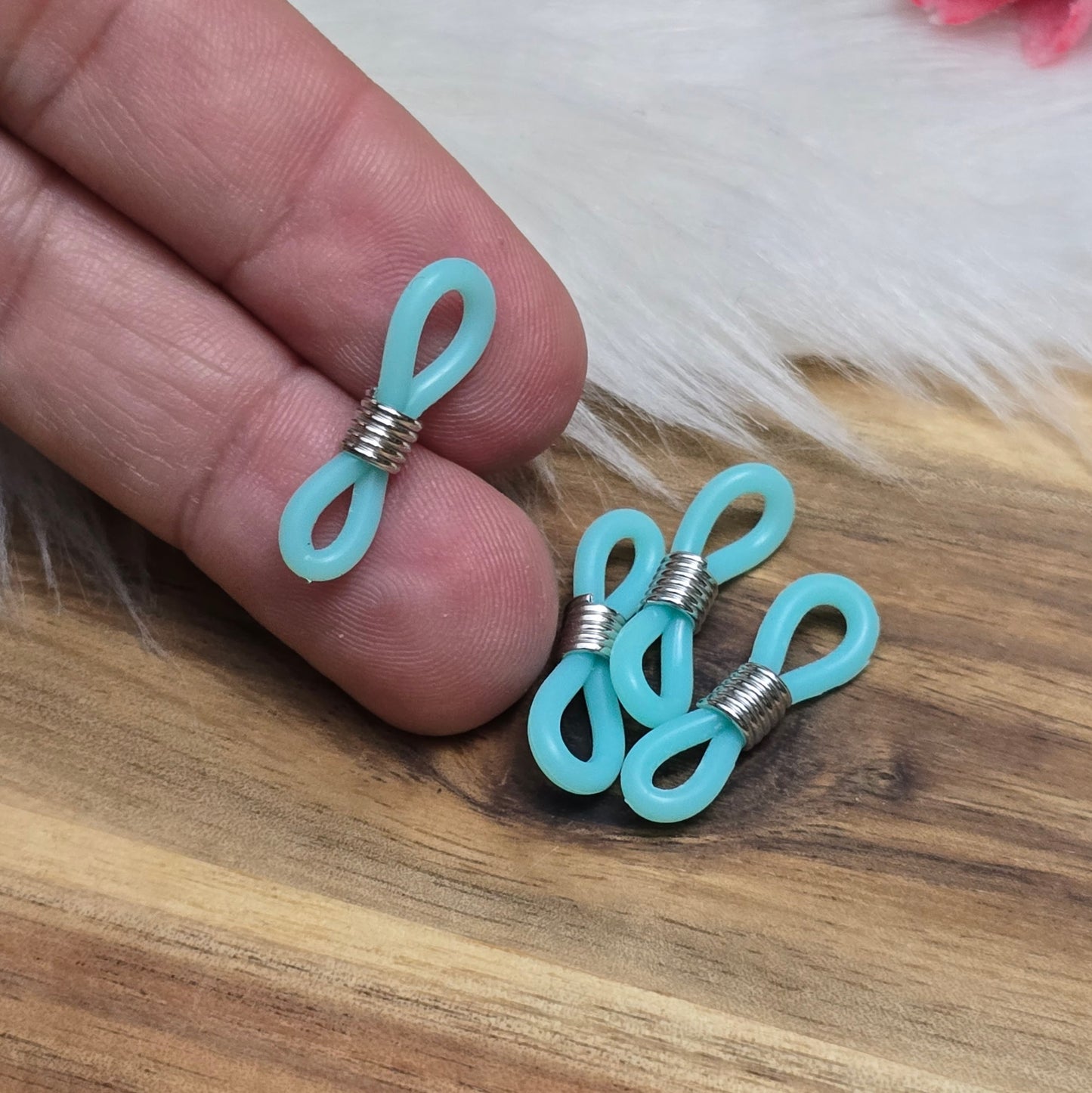 Das Bild zeigt eine Auswahl an Silikonschlaufen für Brillenketten und Maskenhalter in drei Farben: Mint, Rosa und Schwarz. Jede Schlaufe hat eine flexible, weiche Textur und ist so gestaltet, dass sie einfach auf den Bügel der Brille geschoben werden kann. An einem Ende der Schlaufe befindet sich ein kleiner Metallring, der fest angebracht ist, um ein Abrutschen zu verhindern. Die anderen Enden der Schlaufen sind offen, sodass sie mit einem Karabiner oder Biegering leicht an einer Kette befestigt werden kön